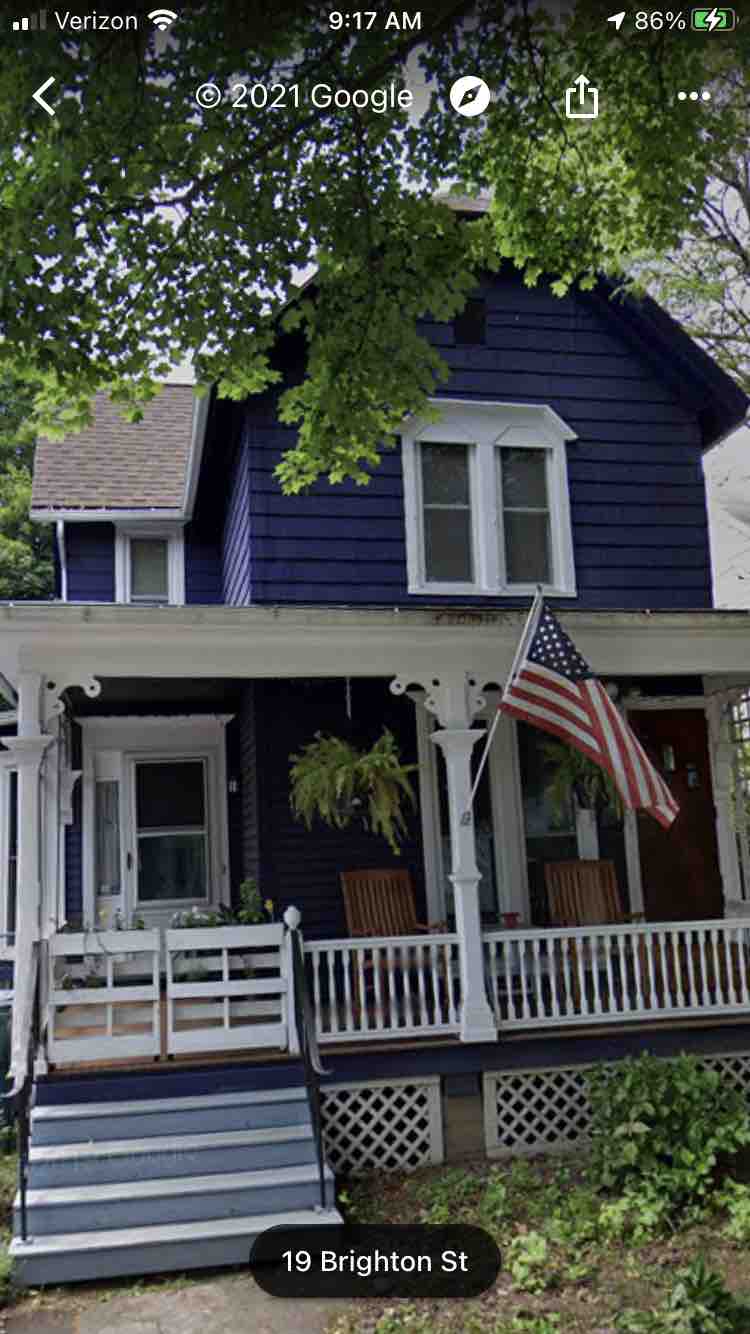 A two-story blue house with white trim is accented by a front porch featuring rocking chairs. An American flag is displayed prominently, while lush greenery frames the entrance. The home is set within a neighborhood, showcasing a classic architectural style.