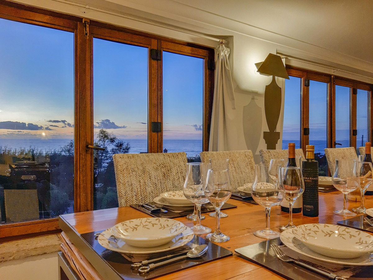 A dining area features a large wooden table set for a meal, showcasing elegant dishware and glassware. Large windows offer a panoramic view of the ocean at sunset, with soft twilight hues illuminating the space. Subtle decor enhances the warm and welcoming environment.