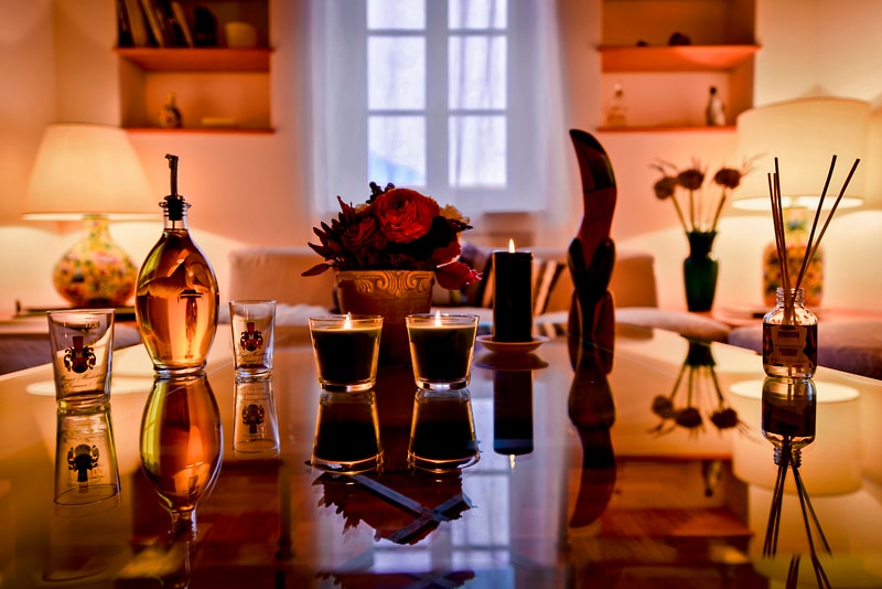A warm living room setting is showcased, featuring a polished table reflecting light from multiple candles and decorative accents. A floral arrangement sits at the center, surrounded by glasses and aromatic diffusers. Soft ambient lighting from lamps enhances the inviting atmosphere.