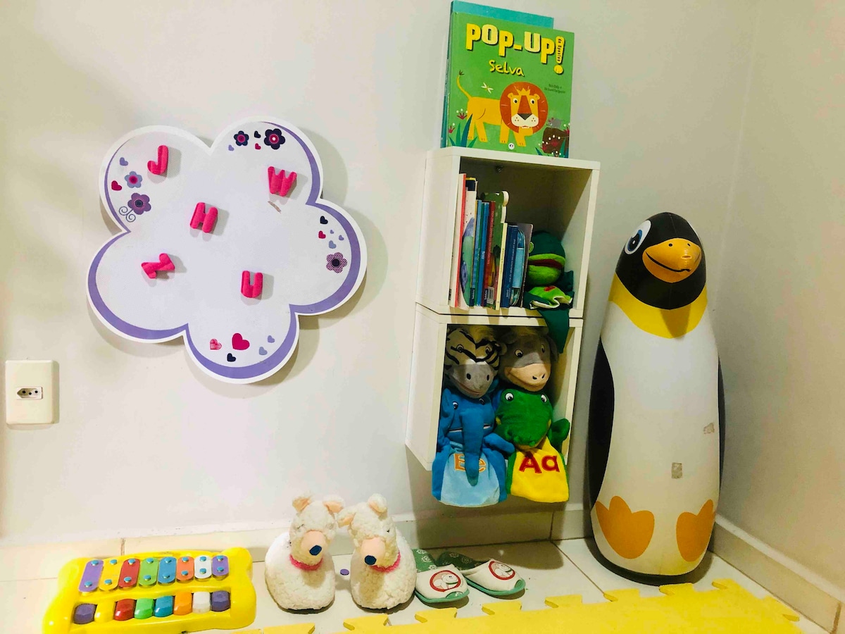 A children's corner is shown, featuring a cloud-shaped whiteboard with colorful magnets. A small bookshelf holds various books and plush toys, including a penguin figure. A playful yellow mat is underneath, alongside a musical toy and two fluffy animal figures.