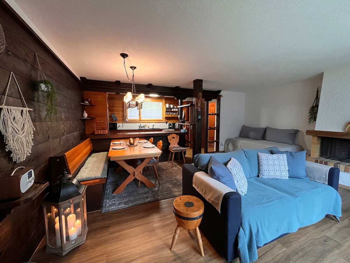 The living area features wooden accents and a cozy ambiance, with a dining table that seats four and a comfortable sofa adorned with cushions. A fully equipped kitchen is visible in the background, and natural light enters through the windows, enhancing the inviting atmosphere.