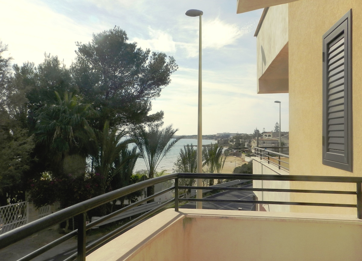 A view from a balcony showcases the distant beach and shoreline. Palm trees line the area, enhancing the natural scenery. The sky is partially cloudy, with soft light illuminating the surroundings. A road can be seen leading to the beach, inviting exploration.