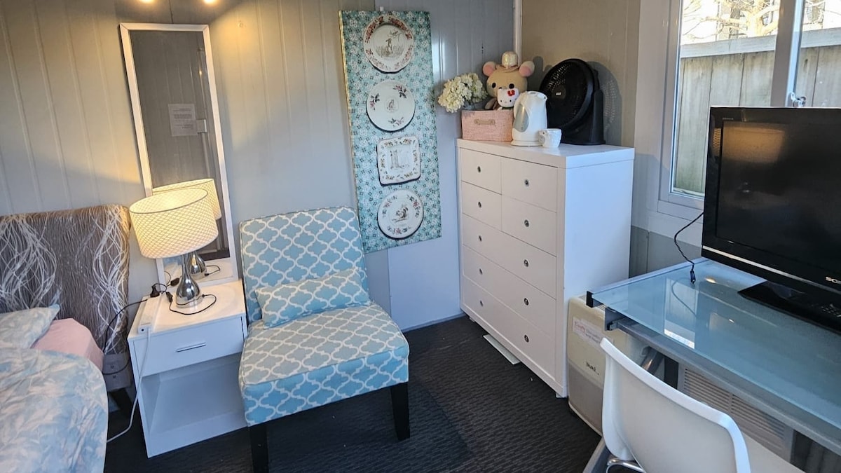 The room contains a queen bed with soft bedding, a bedside table with a lamp, and a cozy accent chair. A dresser provides storage, while a study desk with a chair and a television complete the space. Natural light enters through a window.