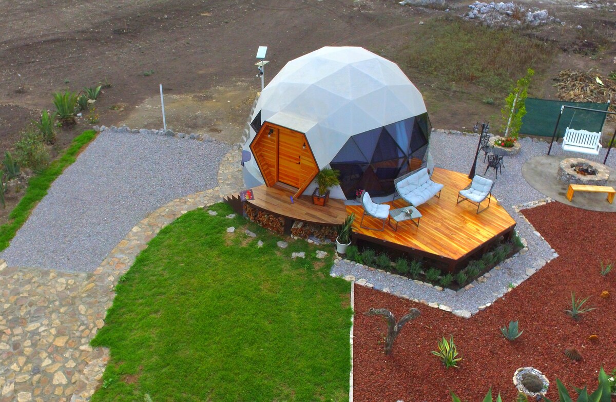 A geodesic dome features a distinctive design set among landscaped grounds. Outdoor seating arrangements provide a comfortable space on a wood deck, surrounded by greenery and decorative stones. Pathways lead to the entrance, enhancing the sense of privacy and tranquility in the area.
