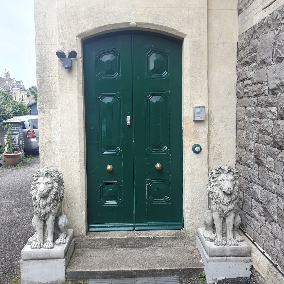 The entryway features a pair of ornate green double doors, flanked by two stone lion sculptures. A security camera is mounted above the door, and a simple doorbell is visible beside the right door. The stone wall adds a textured element to the entrance.