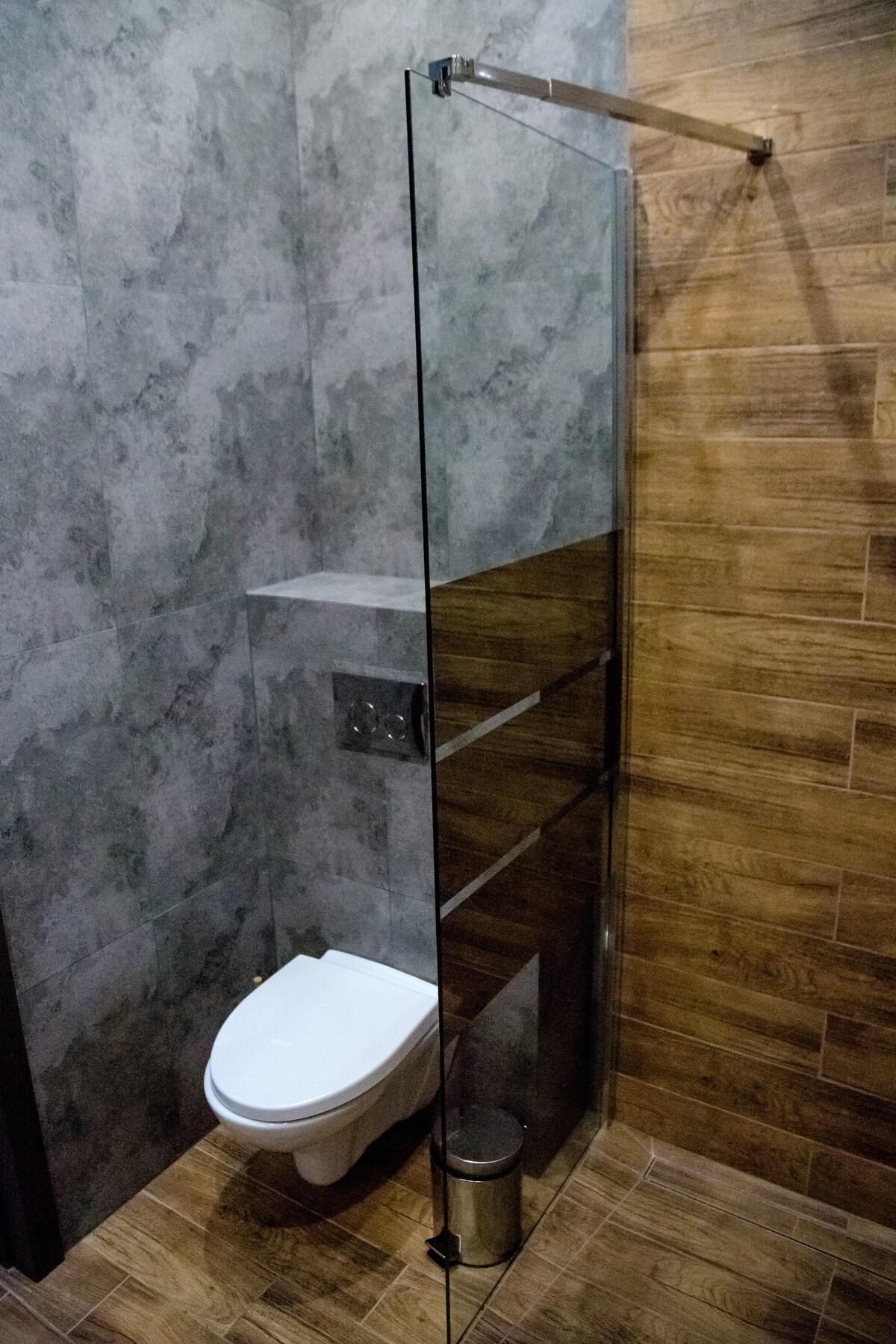 A modern bathroom features a glass shower enclosure with a sleek design. Textured grey and wooden tiles line the walls and floor, while a contemporary toilet is positioned discreetly. A chrome towel bar adds a functional element to the space.