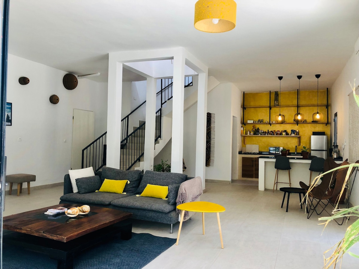 A spacious living area features a comfortable gray sofa with yellow accents, complemented by a low wooden coffee table. The space is illuminated by pendant lighting, with a minimalist kitchen visible in the background. Stairs lead to the upper level, enhancing the open atmosphere.