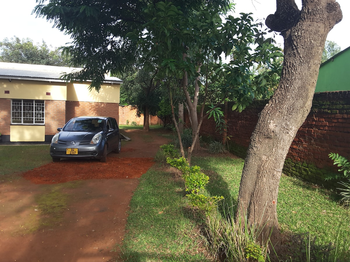 A tree-lined pathway is shown leading towards a house, bordered by green grass and small shrubs. A parked car is visible along the path, with a brick wall marking the property boundaries. The environment appears calm and inviting, surrounded by greenery.