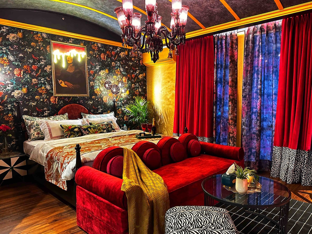 A vibrant bedroom features a plush bed adorned with floral bedding, flanked by rich red curtains. A striking chandelier hangs overhead, while floral wallpaper adds bold character to the space. A red sectional sofa and a glass table with decorative accents complete the cozy atmosphere.
