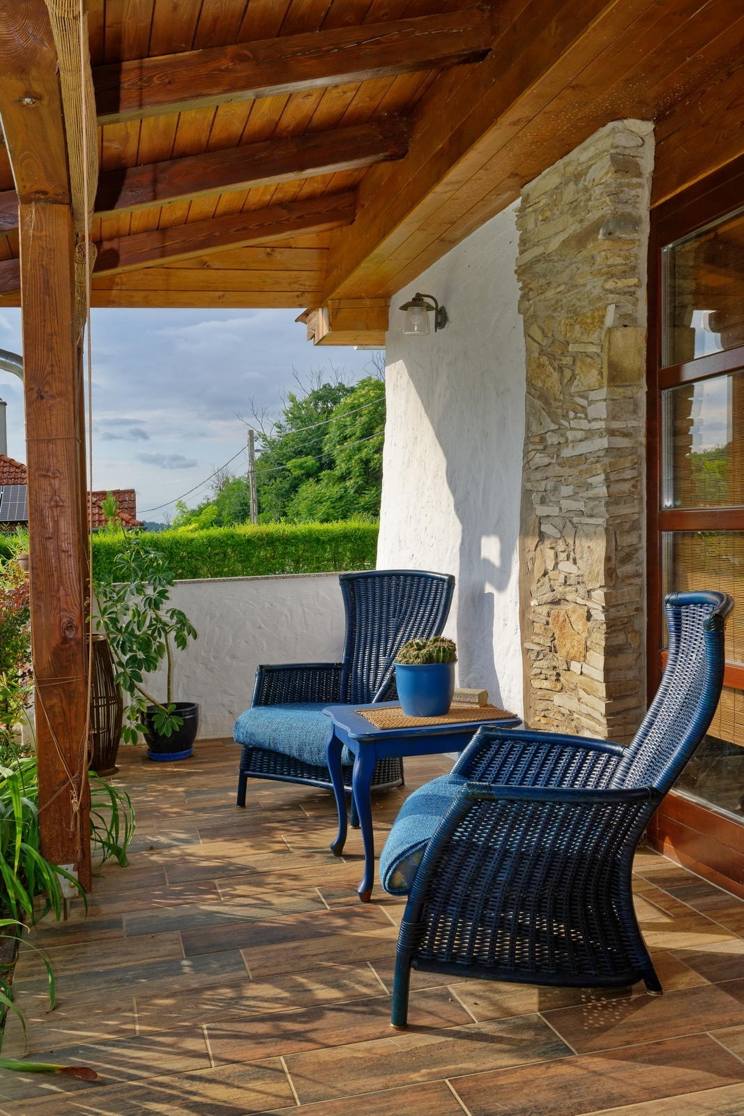 A covered terrace is furnished with two comfortable chairs and a small table, providing a serene outdoor seating area. Lush greenery surrounds the space, with a plant in a blue pot adding a pop of colour. Natural light enhances the welcoming atmosphere.