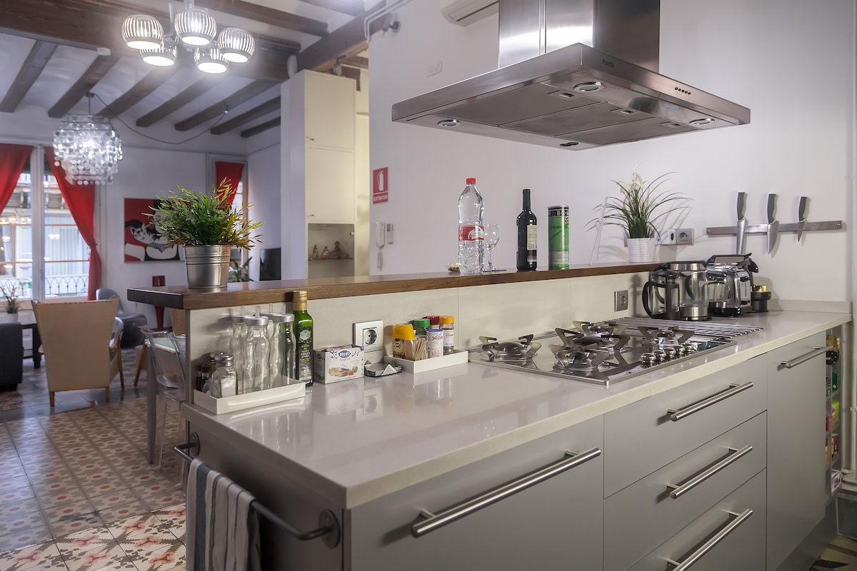 A modern kitchen is displayed with sleek cabinetry and a large island featuring a gas stove. A variety of cooking tools, condiments, and utensils are organized neatly on the counter. Natural light filters through a nearby window, illuminating the space.
