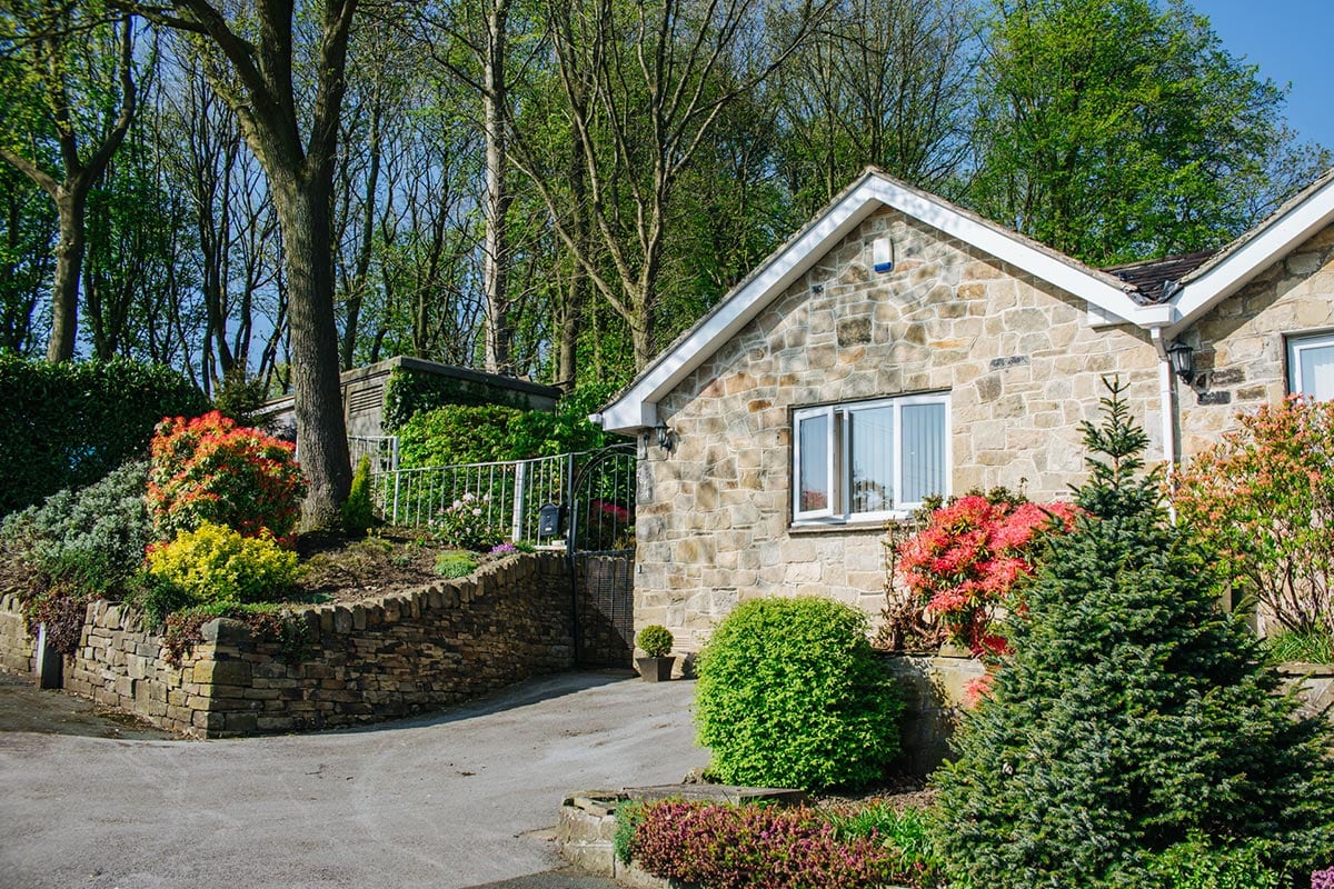 A stone bungalow is surrounded by well-maintained gardens featuring vibrant shrubs and flowering plants. Tall trees provide shade in the backdrop, and a paved driveway leads to the entrance. Natural light is reflected in the windows, enhancing the inviting appearance of the home.
