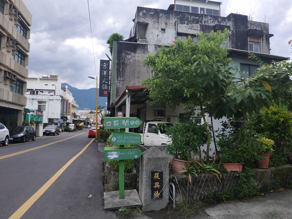 The exterior view features a charming building with outdoor signage and various plants. A signpost with directional arrows indicates nearby attractions, while a small stone monument is visible in the foreground. The street displays a mix of urban architecture and greenery.