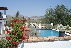 A tranquil scene is presented with a blue pool surrounded by vibrant greenery and flowering plants. The pool area is framed by a traditional fence and offers views of the rolling hills beyond, creating a serene outdoor space.
