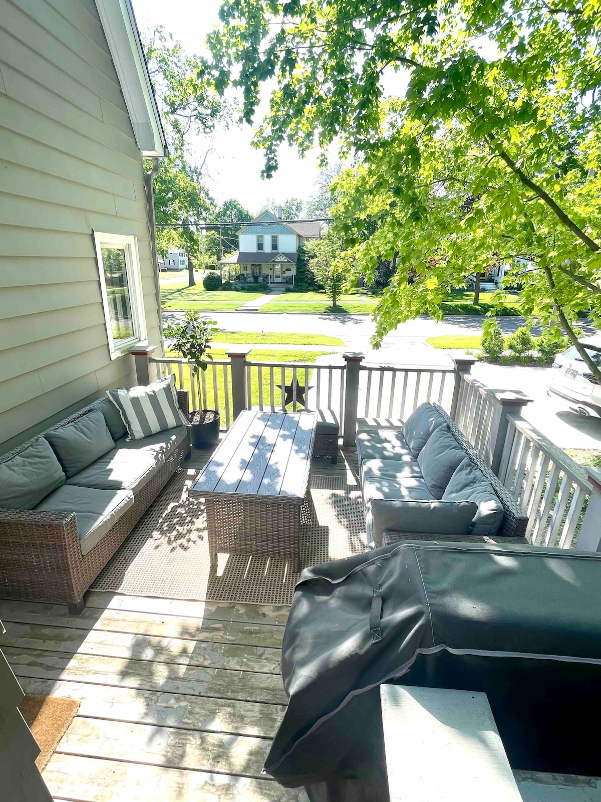 An outdoor seating area is depicted, featuring a gray wicker sofa set and a wooden coffee table. Sunlight filters through the surrounding trees, casting dappled light across the deck. Nearby views of the neighborhood and street are visible.