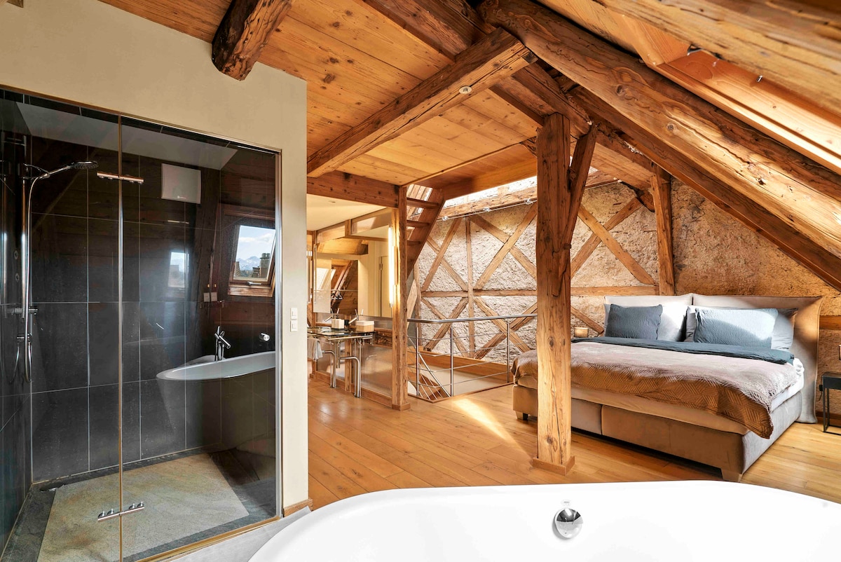 An open sleeping area features rustic wooden beams and warm wood flooring. A spacious bed is complemented by a freestanding bathtub, adding a touch of luxury. A glass shower is present, allowing natural light to illuminate the space through a nearby skylight.