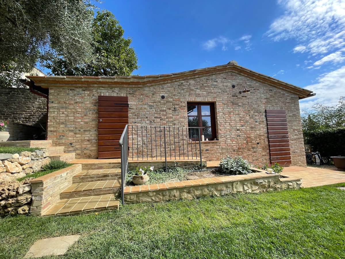 A charming stone structure is framed by well-maintained greenery and steps leading to a welcoming entrance. Two wooden doors are set within the brick façade, complemented by a small garden area filled with plants, creating a serene outdoor setting under a clear blue sky.