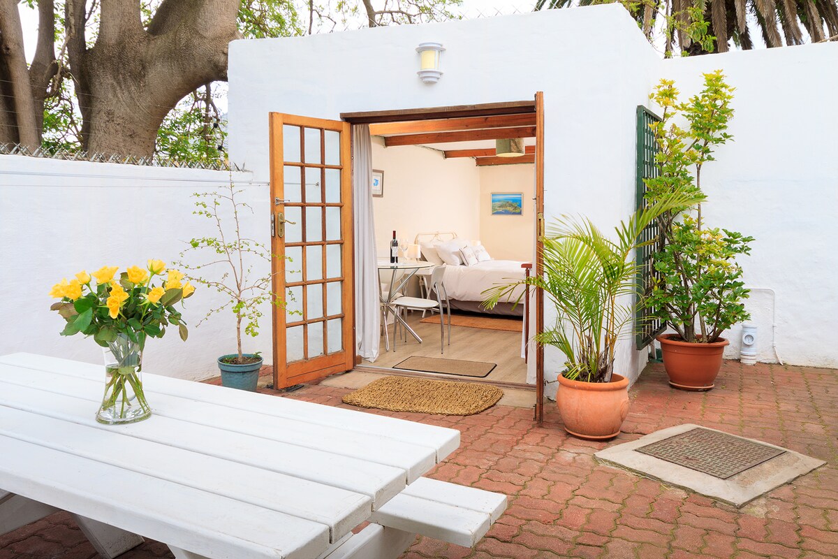 A private outdoor entertaining area is presented, featuring a white picnic table and potted plants, including a yellow flower arrangement. The entrance to the studio is framed by wooden doors, leading to a light and airy interior with a double bed, visible in the background.