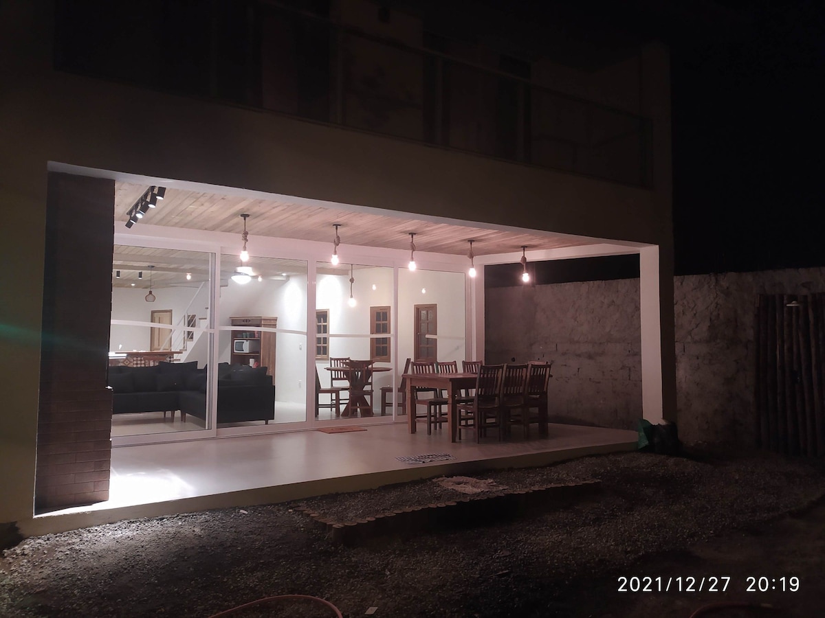 An outdoor dining area is illuminated by pendant lights, featuring a spacious table surrounded by wooden chairs. The interior space is visible through large glass doors, revealing a comfortable living area. A textured wall adds depth to the setting, enhancing the overall ambiance.