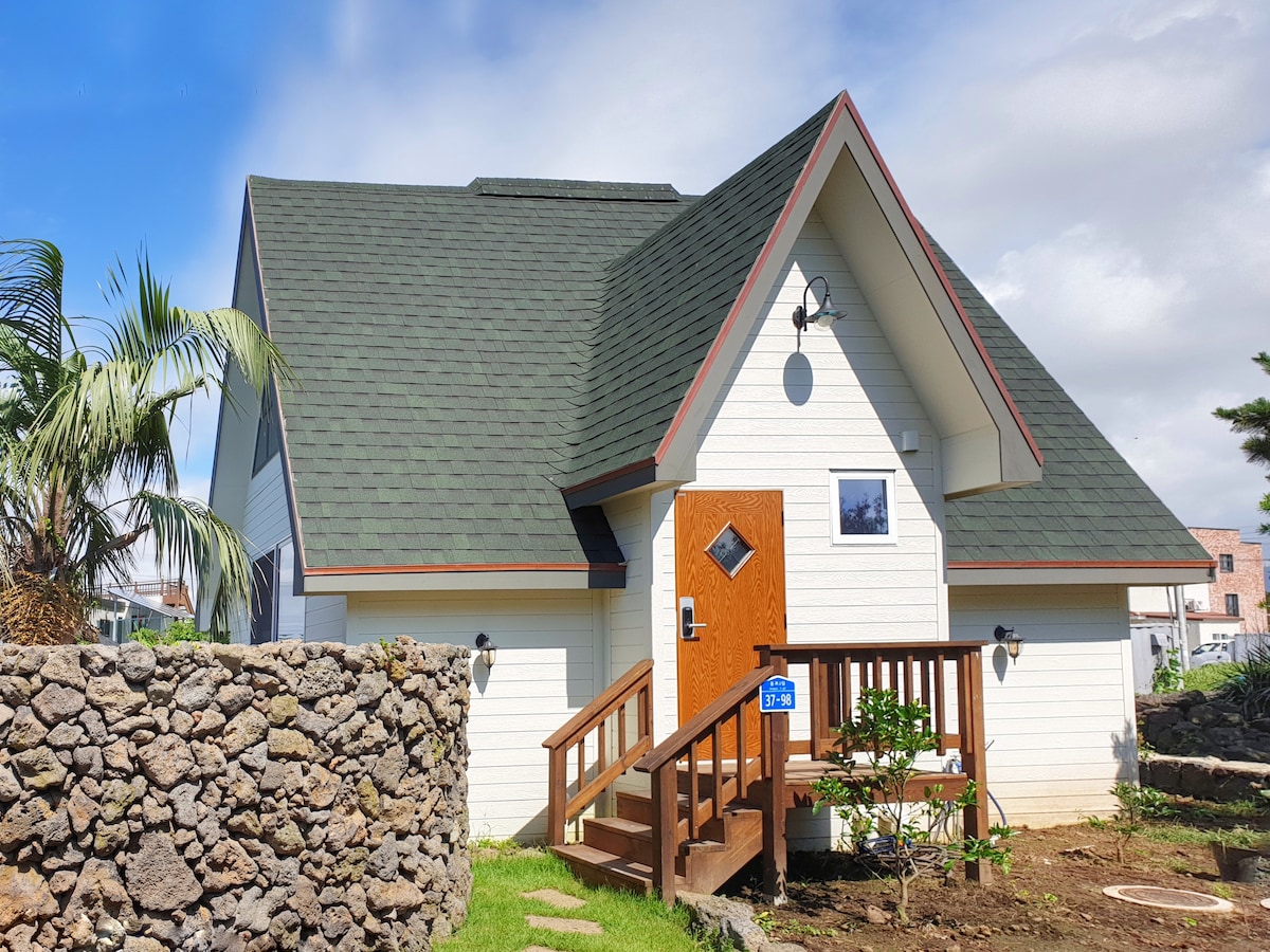 A charming two-story cottage is showcased, featuring a steeply pitched roof and a wooden front door. Lush greenery and a stone wall add to the inviting exterior. A small garden is visible in the foreground, enhancing the overall tranquil setting.
