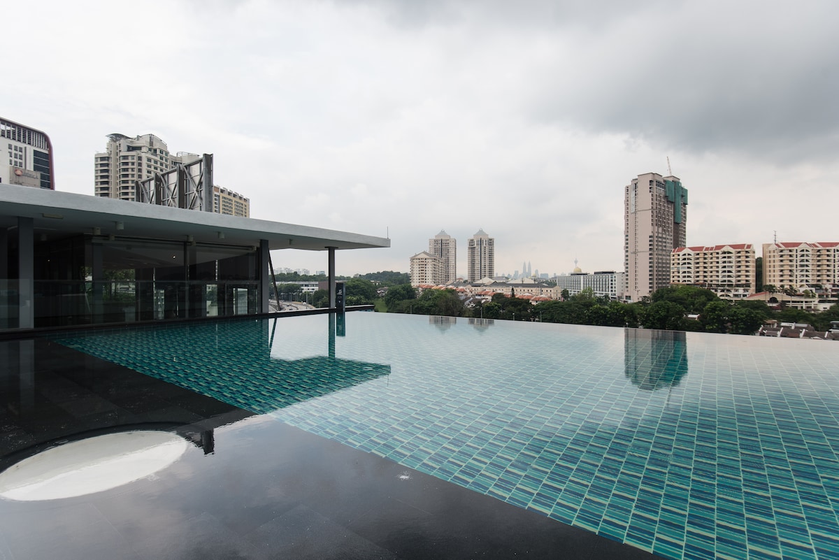 An infinity pool is displayed against a backdrop of urban high-rises, with water reflecting the cloudy sky. The pool area is designed with blue and green tiles, enhancing the overall aesthetic of the outdoor space.