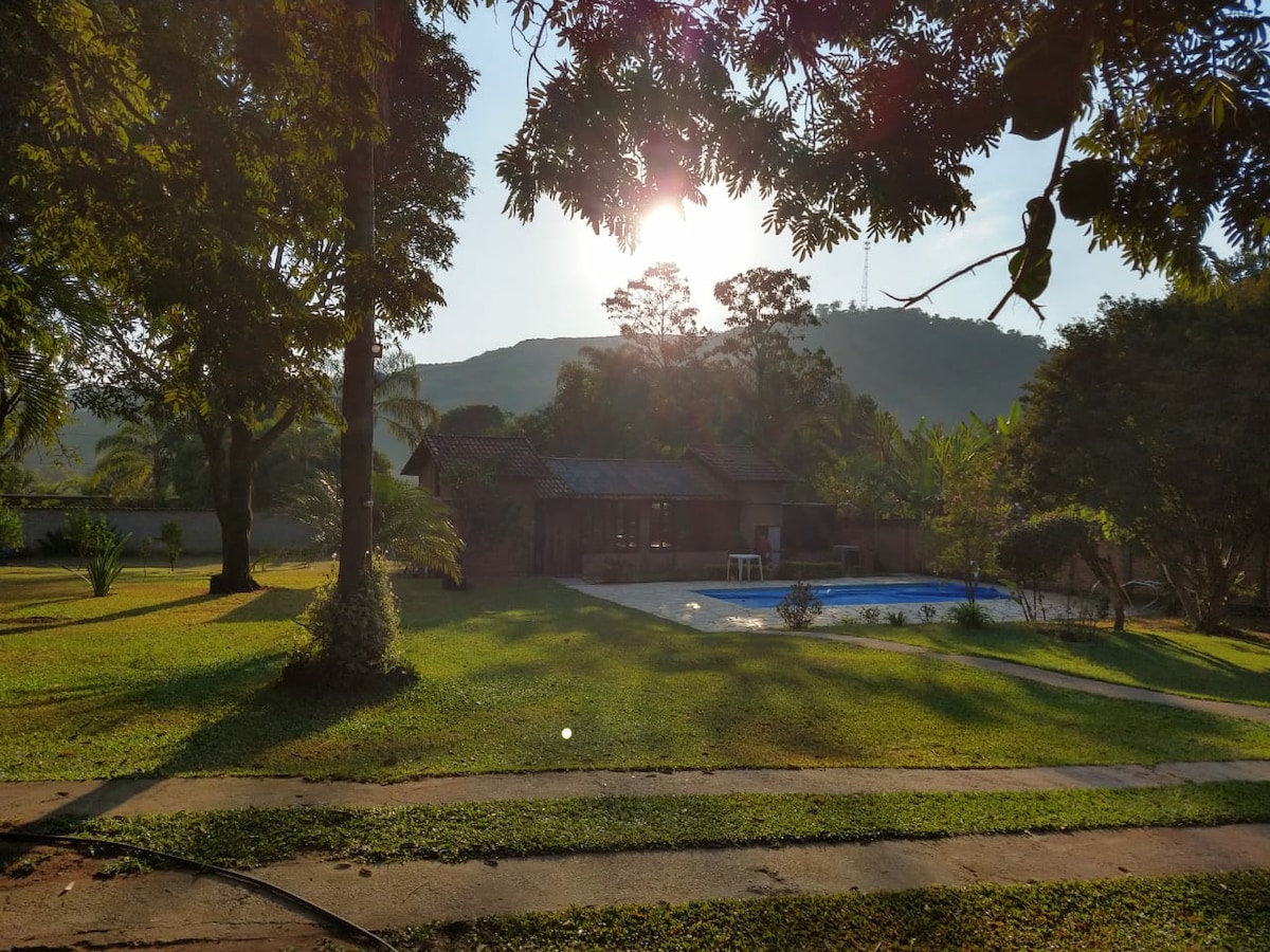 The serene outdoor area features a spacious lawn framed by lush trees, with sunlight filtering through the leaves. A pool is visible in the background, complemented by mountains rising in the distance, creating a tranquil setting for relaxation.