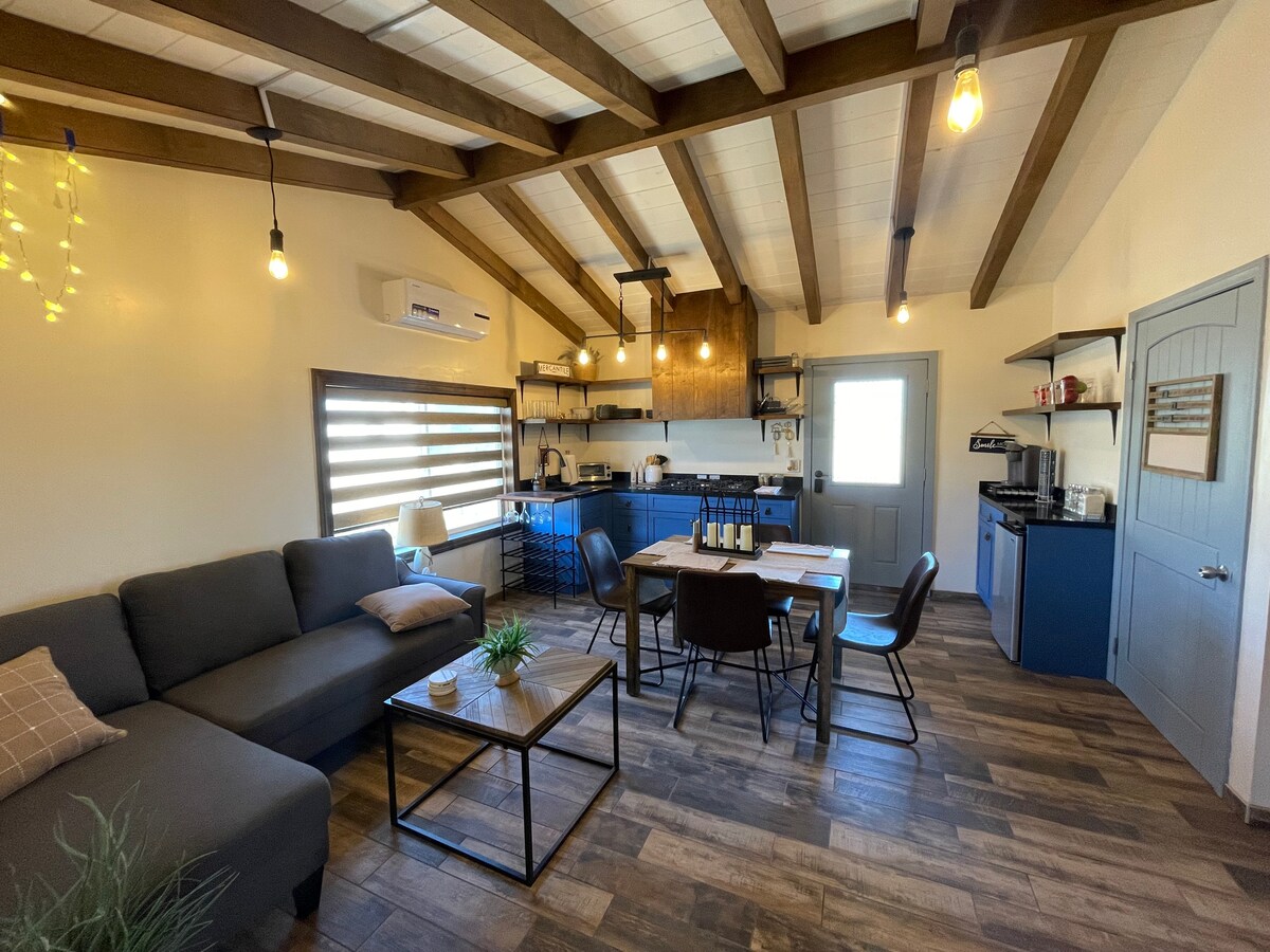 The spacious living area features exposed wooden beams and a cozy seating arrangement with a dark gray sofa. A dining table is situated near a well-equipped kitchen, showcasing blue cabinetry and modern appliances. Light fixtures hang from the ceiling, adding a warm ambiance.