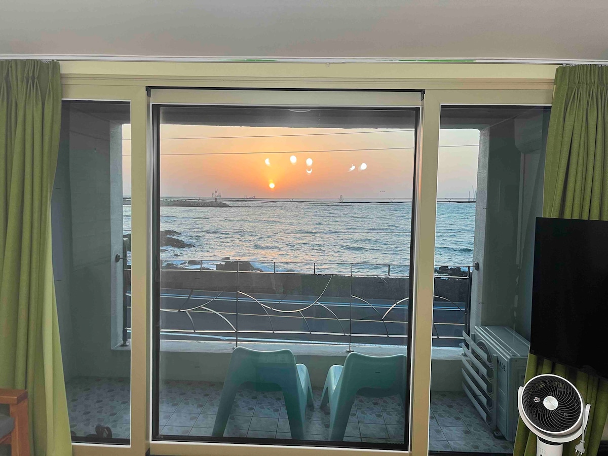 Large glass windows frame a stunning sunset over the ocean, with a serene view of the water stretching to the horizon. Two turquoise chairs are positioned by the window, inviting relaxation while enjoying the beautiful scenery.