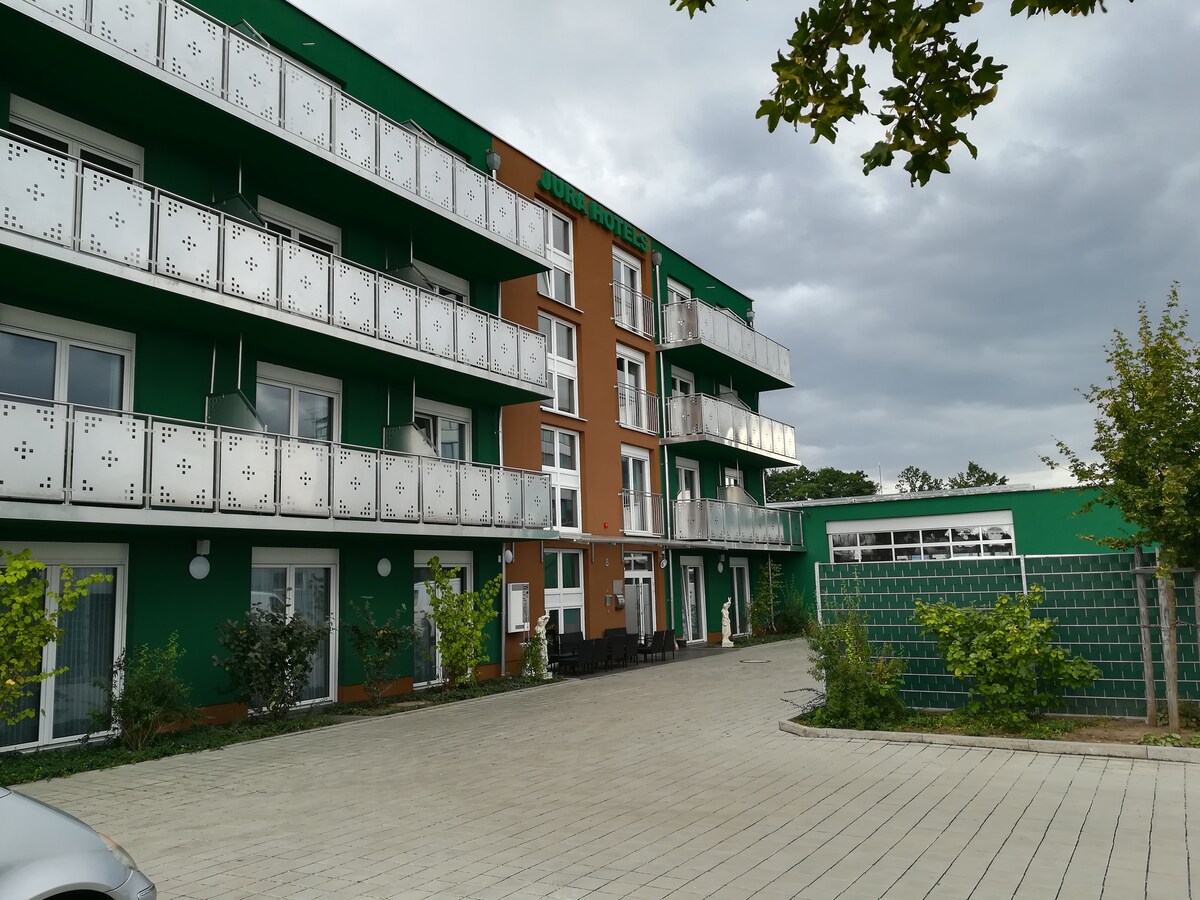 The exterior of a modern hotel is visible, featuring multiple levels with balconies. The building is painted in vibrant green and earthy tones. A paved area leads to the entrance, with landscaped greenery and seating arrangements positioned nearby.