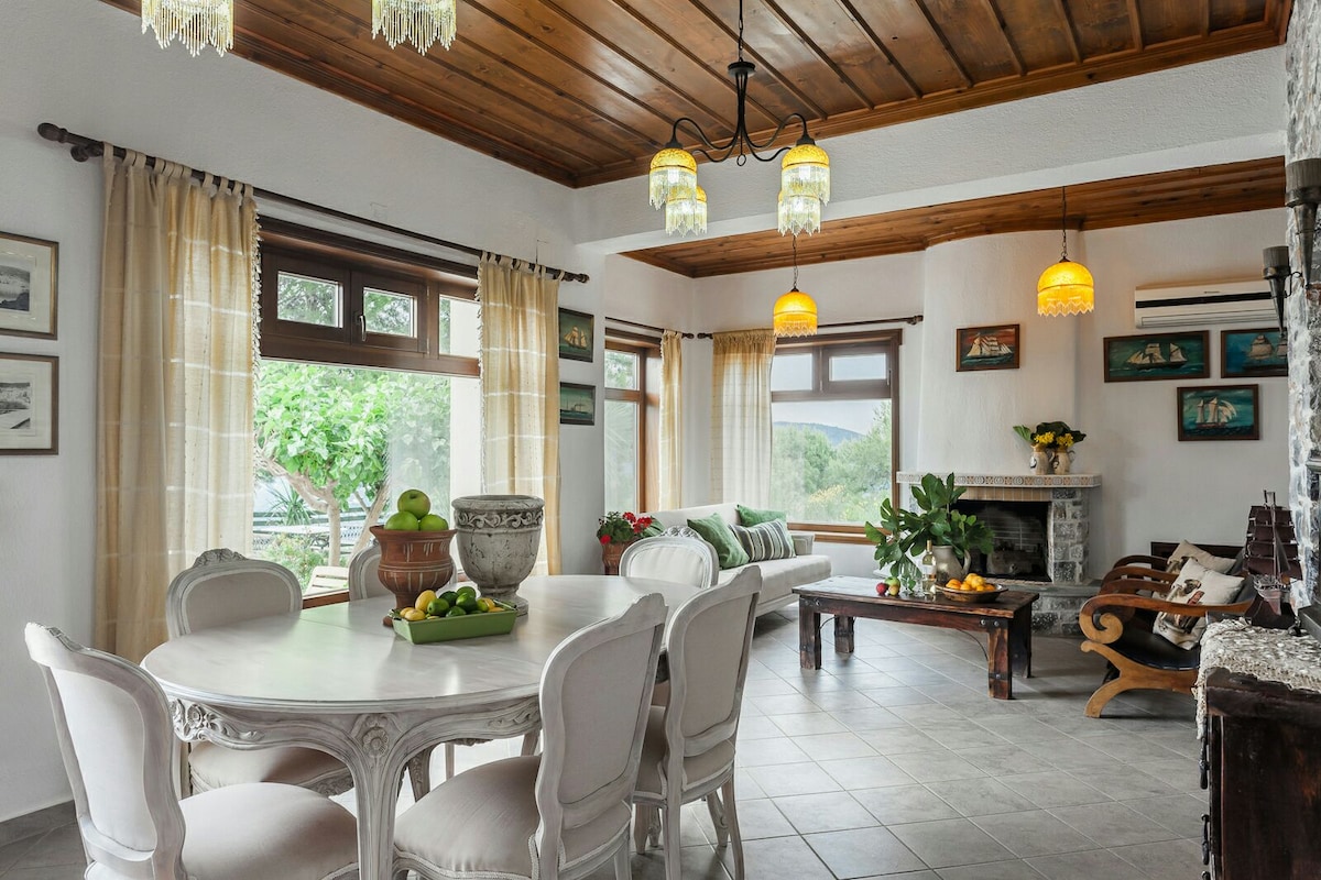 An inviting dining area features a round table surrounded by upholstered chairs. Natural light filters through large windows, highlighting a cozy seating space with a plush sofa and decorative plants. A classic chandelier and warm-toned lamps add to the ambiance.