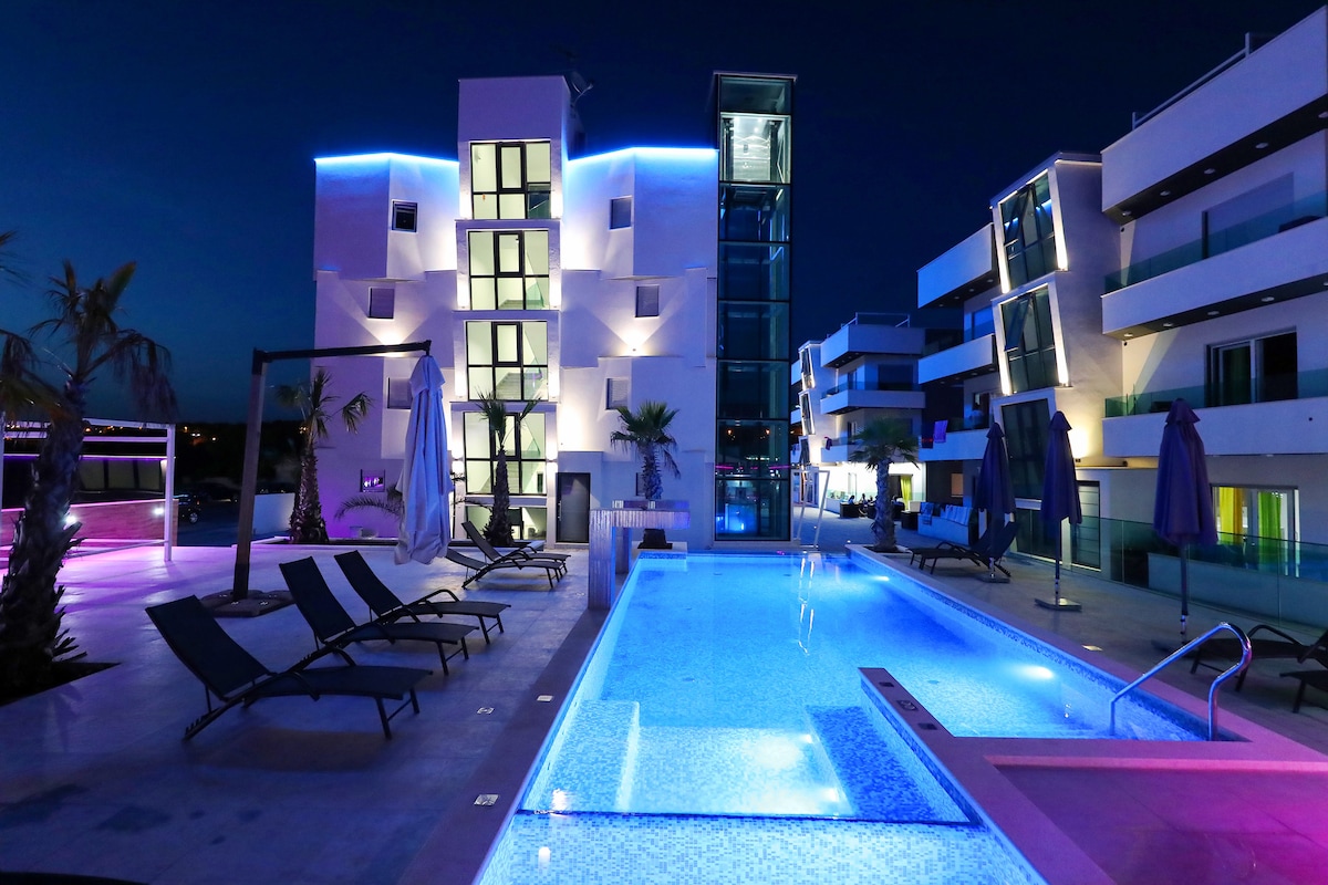 An illuminated outdoor area features a modern building with sleek lines, surrounded by lounge chairs. A swimming pool with varying shades of blue and purple light reflects the night sky. Nearby, palm trees add a touch of greenery, enhancing the contemporary setting.