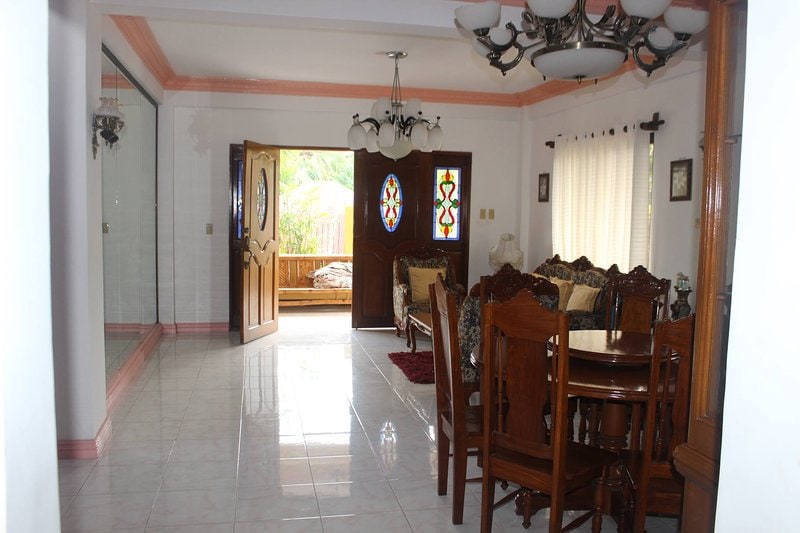 The image shows a bright living area featuring a dining table and chairs made of dark wood. A spacious seating arrangement, decorated with intricate woodwork, is positioned against the wall. Large glass doors lead to an outdoor area, allowing natural light to fill the room.