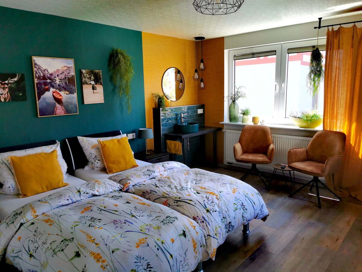 A modern guest room is designed with vibrant green and yellow walls. Two spacious beds are adorned with floral bedding. A round mirror and decorative plants enhance the aesthetic, while large windows allow natural light to illuminate the area, complemented by soft curtains.