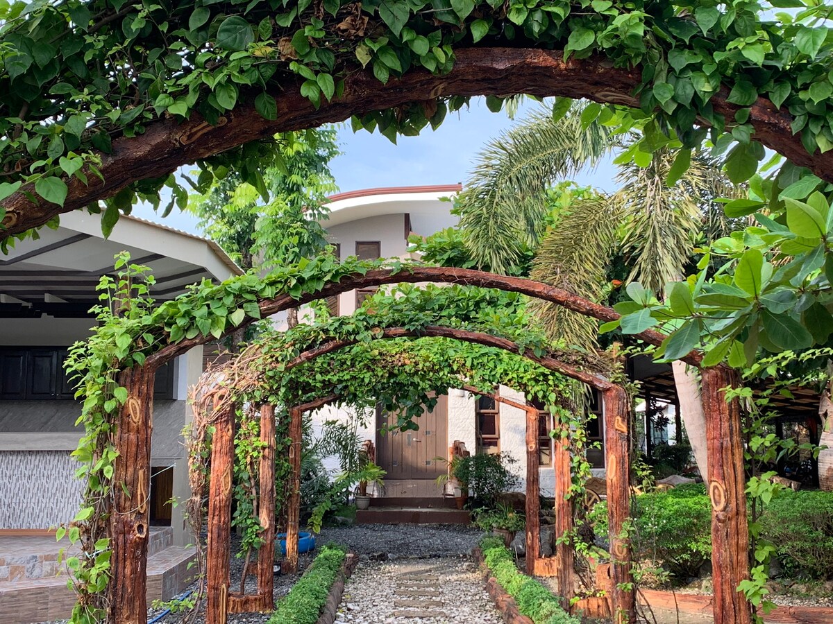 An entrance pathway is framed by wooden arches covered with lush greenery, leading towards a building surrounded by various plants. The neatly arranged stones create a defined walkway, contributing to the serene atmosphere of the outdoor space.