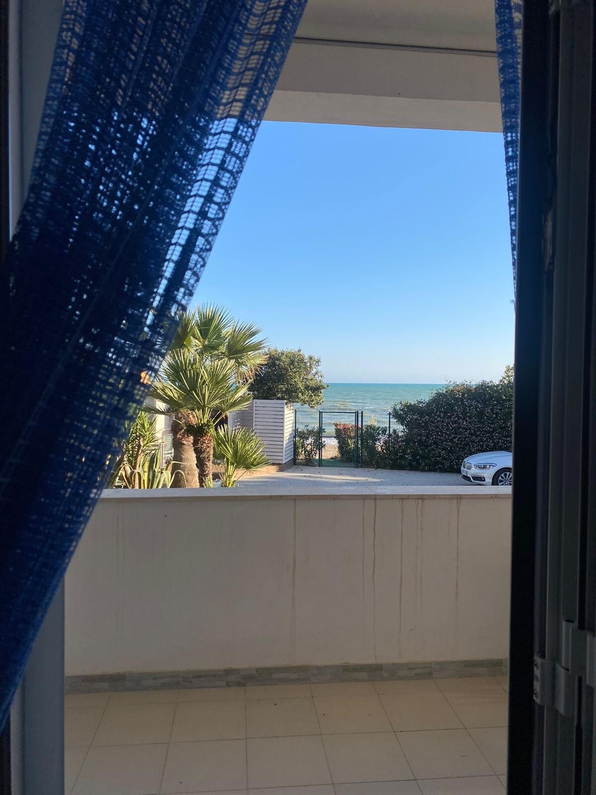 A view from the villa's veranda reveals a clear blue sky and the sparkling sea. Tropical plants are visible on the left, while a pathway leads to the beach. A parked car is positioned just beyond the lush greenery.