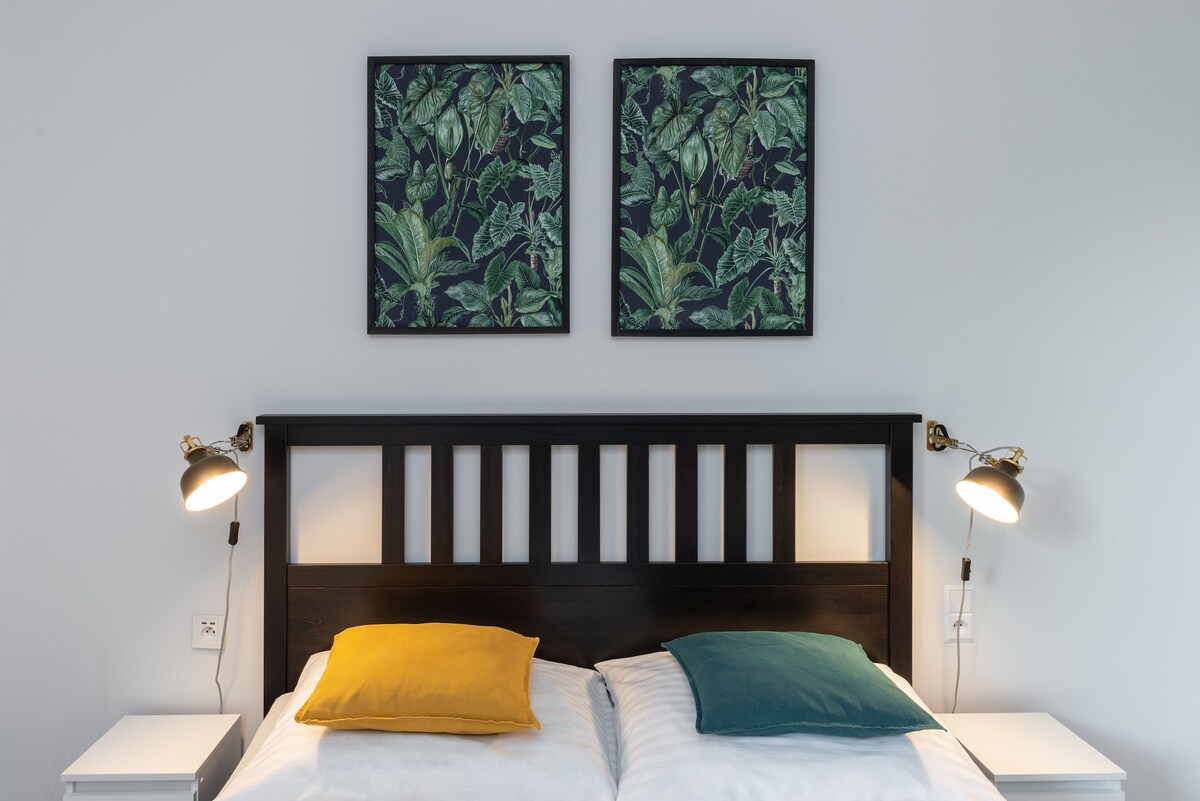 A double bed is presented with white linens and two decorative pillows in yellow and teal. Above the bed, two framed artworks featuring botanical designs are displayed. Simple bedside lamps are positioned on each side, illuminating the sleeping area.