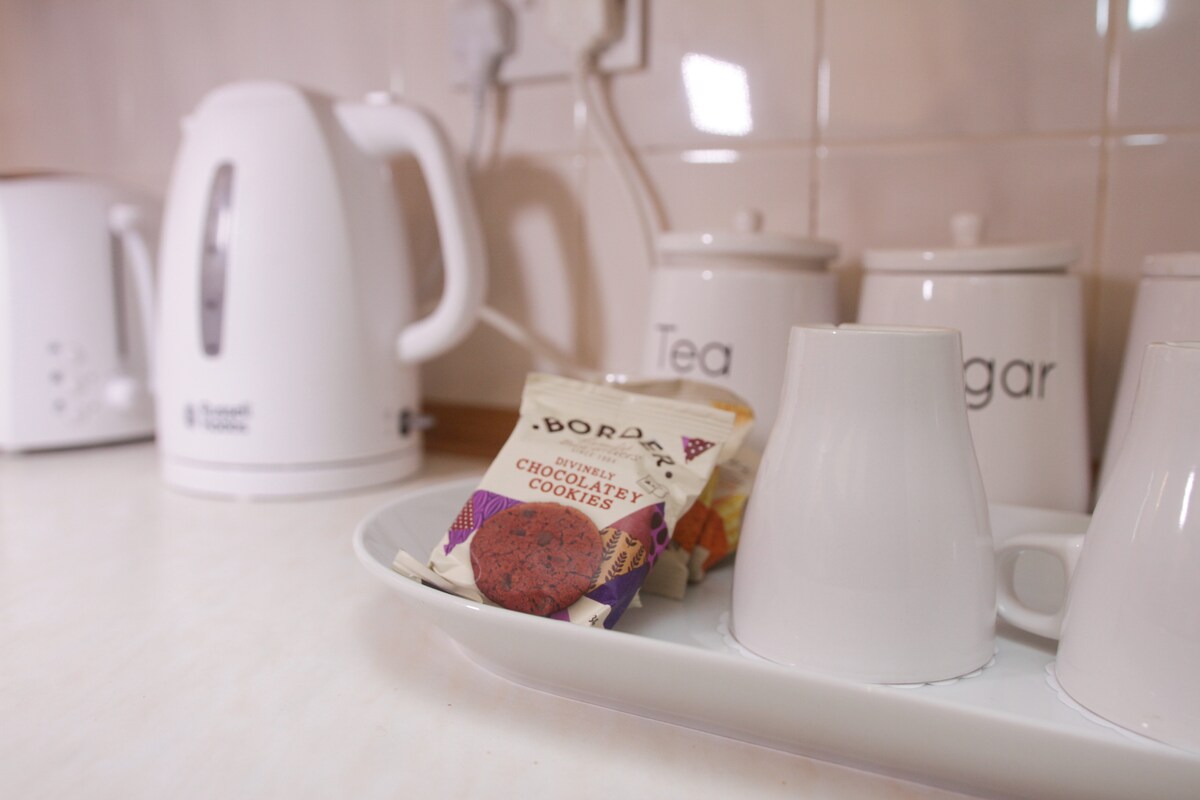 A clean kitchen countertop features a white kettle and storage jars labeled for tea and sugar. Two white cups are situated on a tray, alongside a package of chocolate cookies. Natural light enhances the bright, inviting atmosphere of the space.