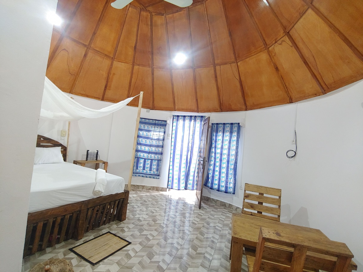The interior features a circular design with a wooden ceiling accentuated by natural light from the entrance. A spacious bed is placed with a mosquito net above, complemented by a rustic wooden table and chairs nearby. Blue-and-white striped curtains frame the doors.