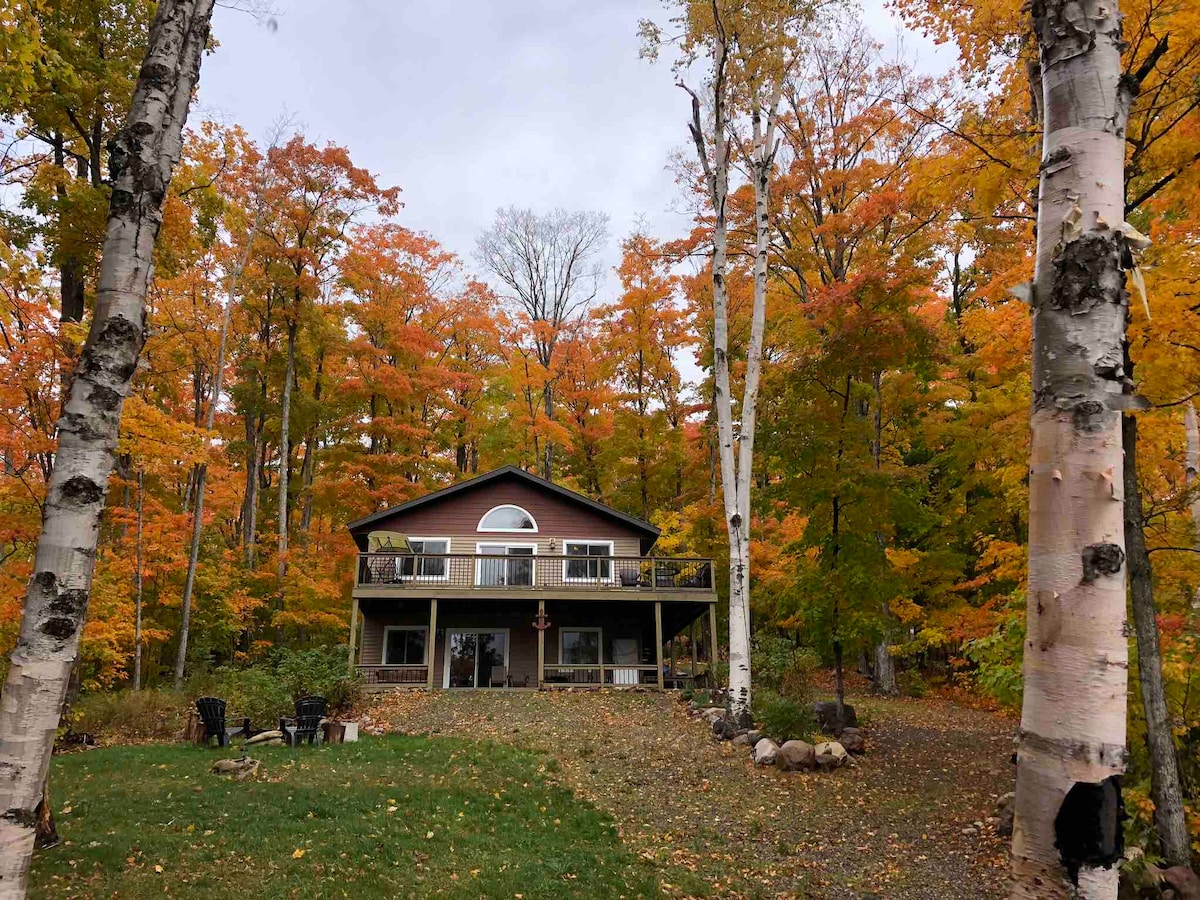A cozy cottage is surrounded by vibrant autumn foliage, showcasing a mix of orange and yellow leaves. The structure features a large wrap-around deck with multiple entry points and large windows, providing a warm feel against the backdrop of tall white birch trees.