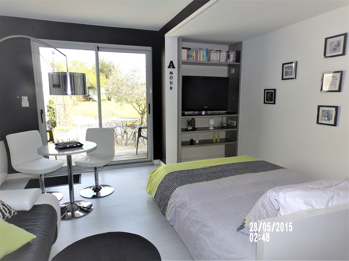 A modern living space features a spacious bed with a gray and green bedspread. Natural light filters through large sliding glass doors, which open to an outdoor area. A circular dining table with two white chairs and a black sofa are arranged thoughtfully within the room.