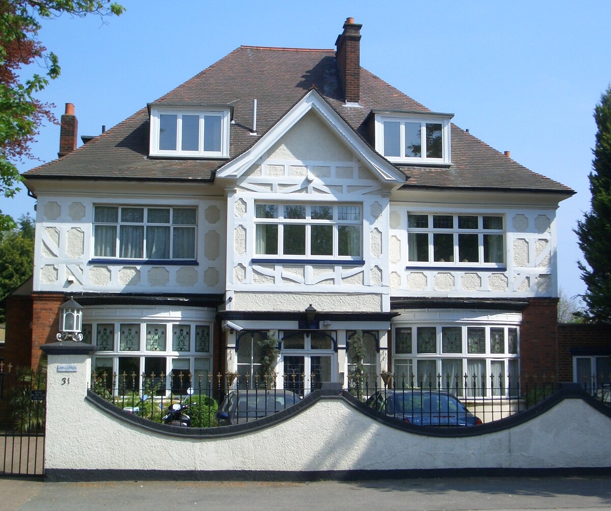 The exterior of a large, three-story Edwardian house features distinct white and light-colored detailing. Multiple large windows illuminate the front facade, with a welcoming entrance framed by an elegant arch. A well-maintained garden and decorative fence enhance the property’s appeal.