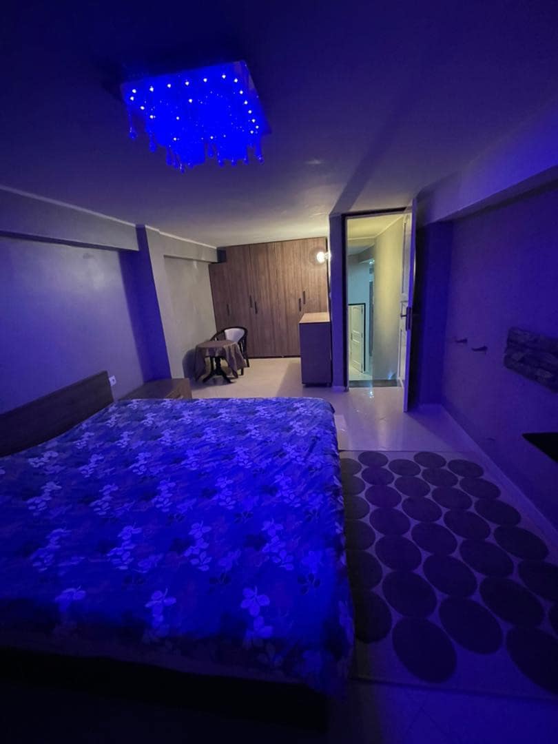 A dimly lit bedroom features a spacious bed covered with a floral-patterned blanket. A unique blue chandelier casts soft illumination. A rug with a circular design adds texture to the tiled floor, while cabinets and a chair are visible in the background.