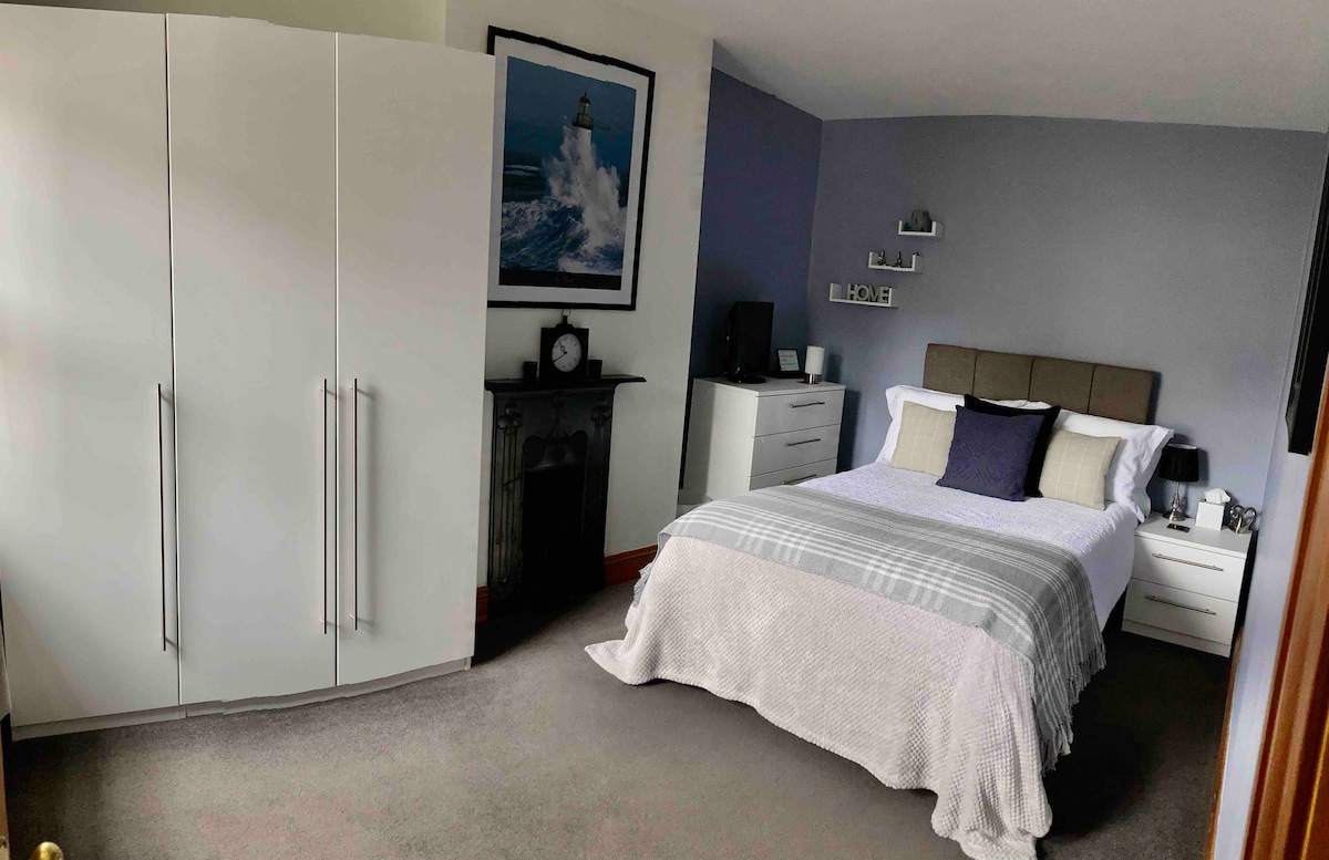 A modern double room is presented, featuring a spacious bed adorned with neutral bedding. A wardrobe and bedside tables provide ample storage, while a decorative fireplace adds character. Artwork of a lighthouse hangs on the wall, complementing the soothing grey tones of the room.