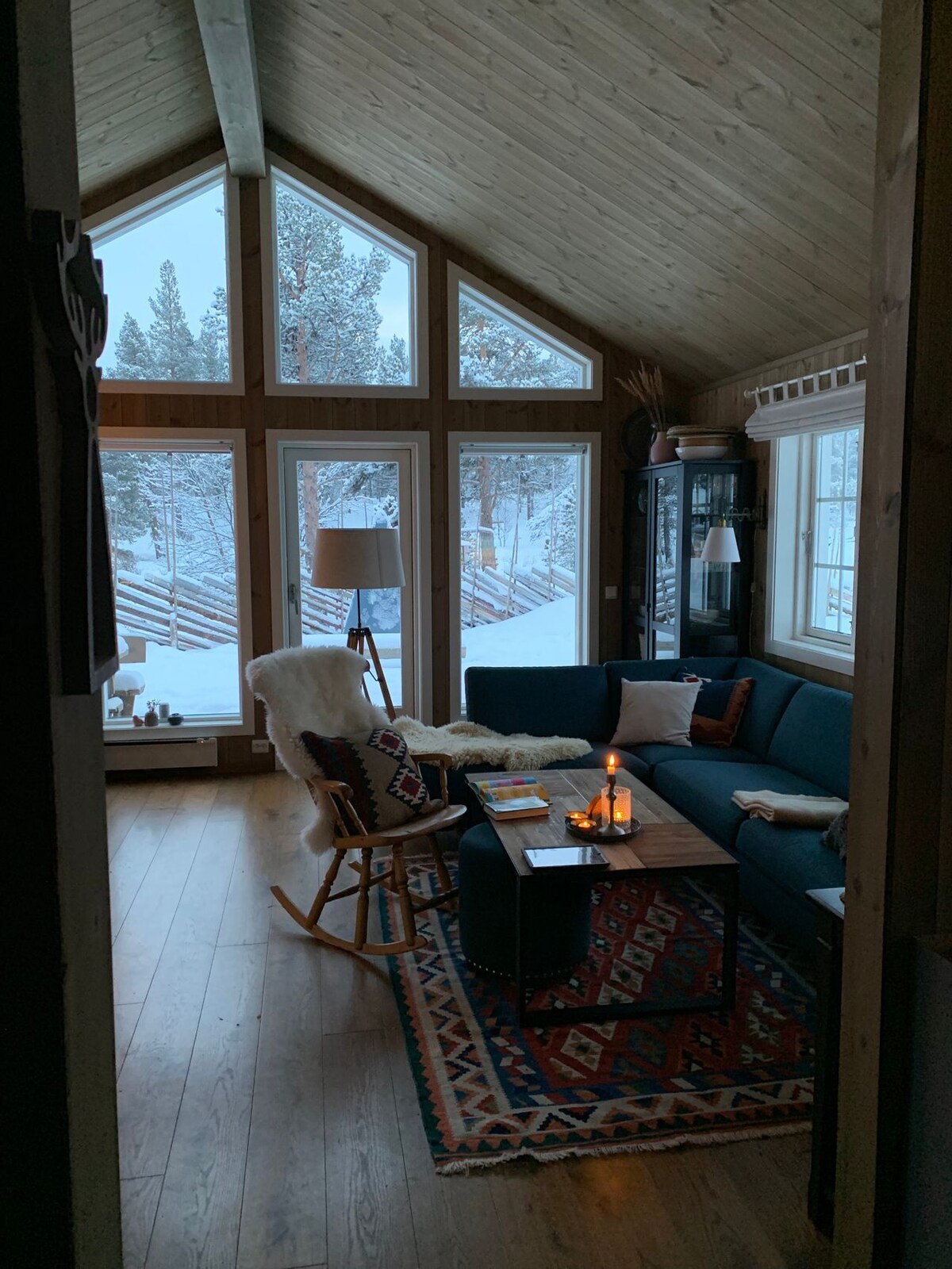 The living area features tall windows allowing natural light, with snow-covered trees visible outside. A blue sofa is positioned beside a wooden coffee table, which holds a candle and magazines. A rocking chair and patterned rug contribute to the cozy ambiance.