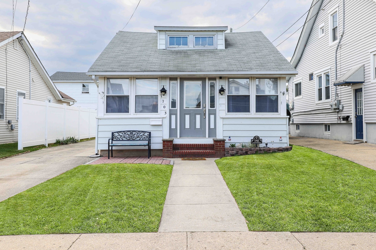 A vintage bungalow is displayed from the front, featuring a gray roof and white siding. The welcoming entry is accentuated by two front doors. A small black bench sits on a red brick patio, surrounded by a manicured green lawn, while the landscape is defined by neat walkways.