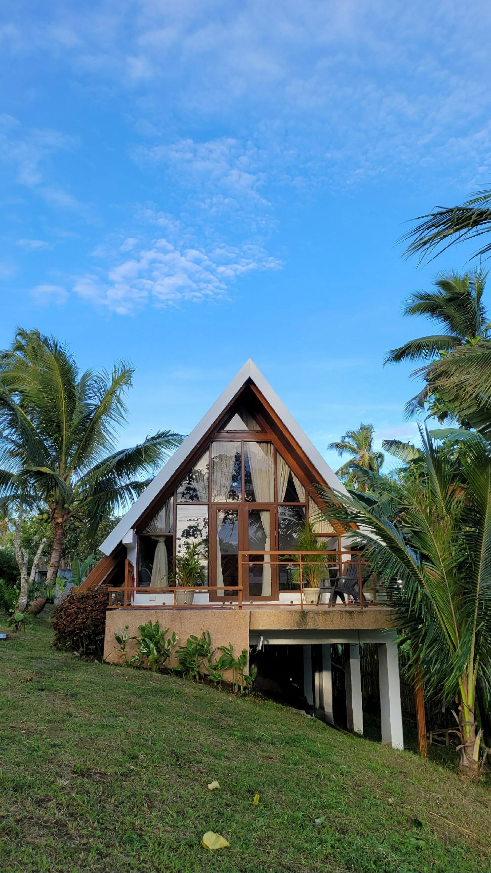 The charming triangular structure features large glass windows that allow ample natural light. Surrounded by palm trees and greenery, the home is set on a gently sloped lawn, enhancing its cozy, welcoming appearance against a clear blue sky.