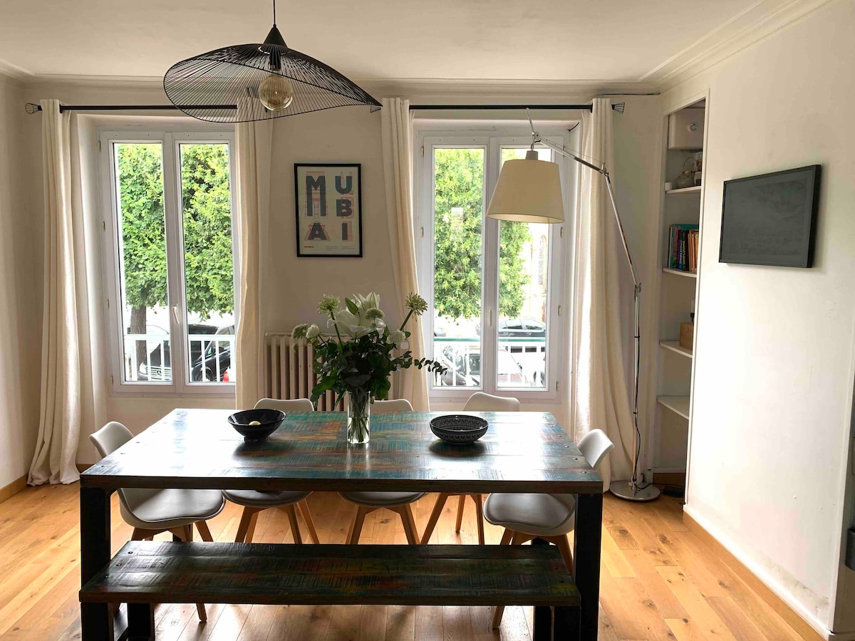 A dining area features a wooden table surrounded by several chairs. Natural light enters through two large windows adorned with light curtains. A decorative vase with white flowers sits at the center of the table, while a floor lamp is positioned beside it.
