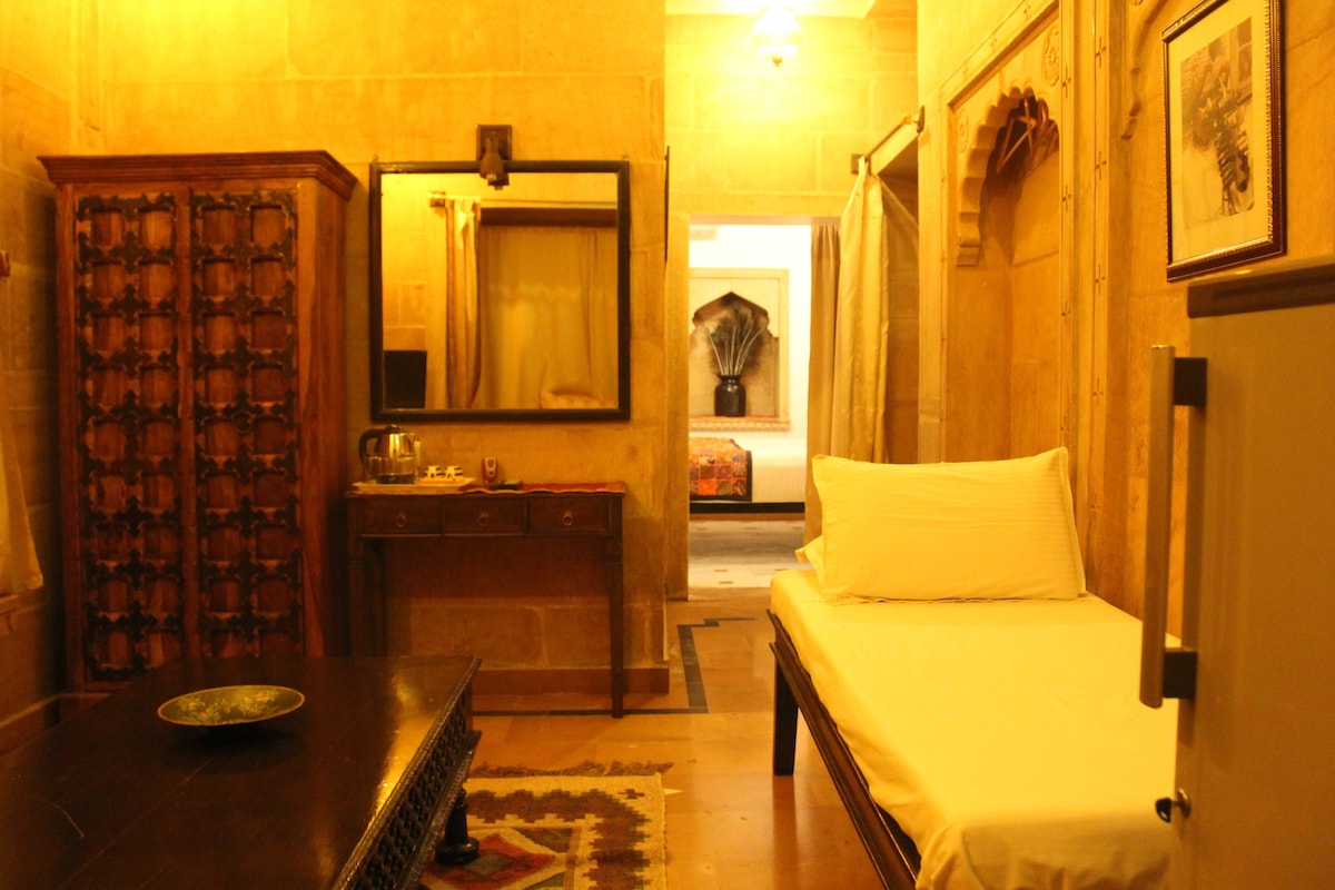 A traditional room features textured sandy walls and antique furnishings, including a wooden wardrobe and side table. A comfortable single bed is positioned alongside a decorative table, while a large mirror reflects a cozy ambiance. Soft lighting casts a warm glow throughout the space.