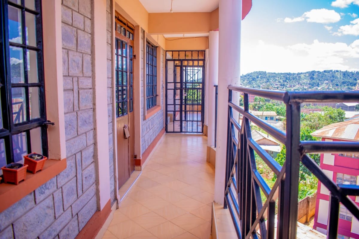 A spacious balcony area features a mix of stone and painted walls, providing an inviting corridor. Large windows enhance natural lighting, offering views of the surrounding landscape. Potted plants add a touch of greenery to the space, while the railing ensures safety and style.