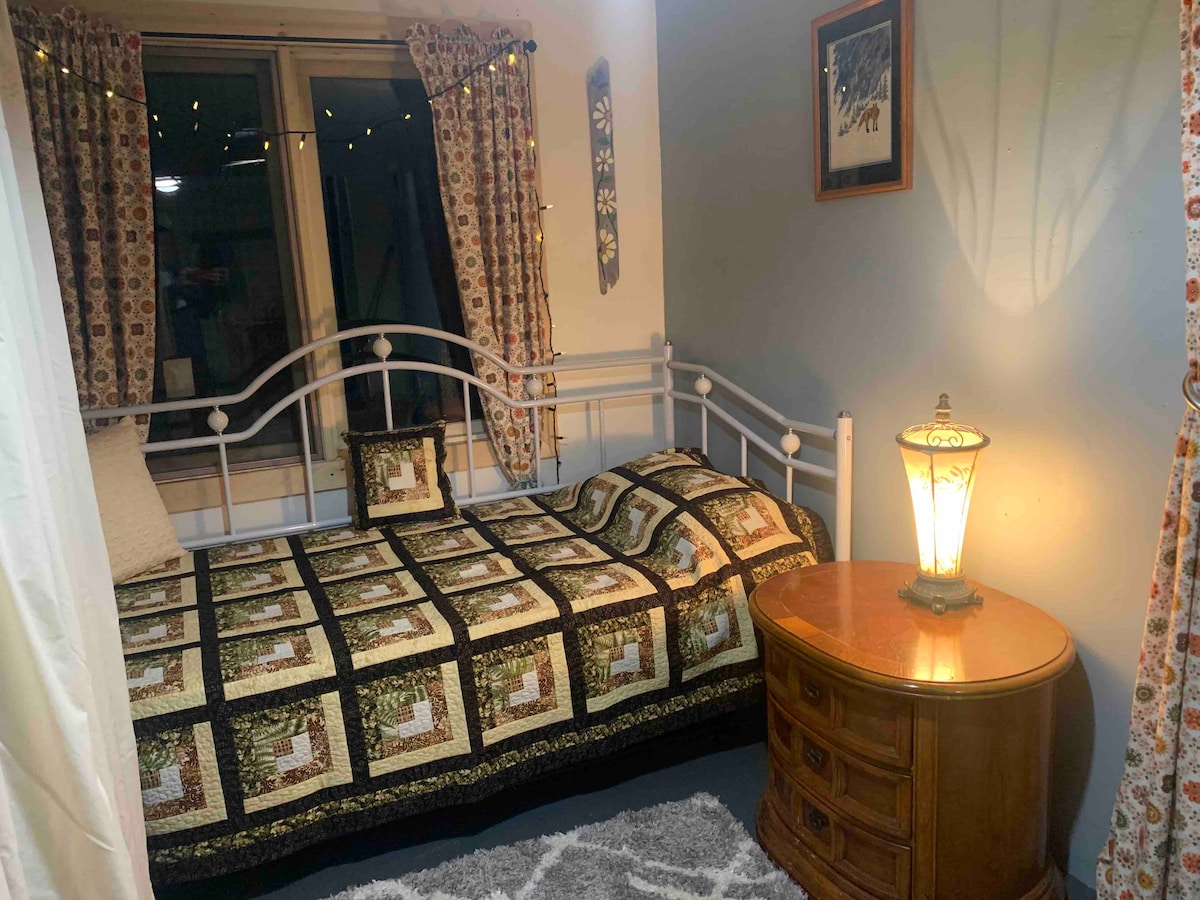 A sleeping area is presented with a metal frame bed covered in a patterned quilt featuring earthy tones. A wooden bedside table and a traditional lamp provide soft lighting, while a window with patterned curtains allows for natural light. Decorative accents include wall art and a cozy rug.
